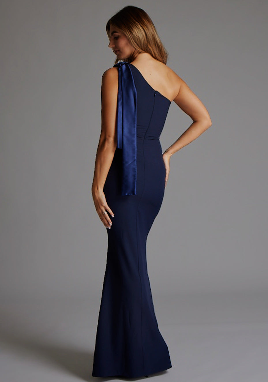 Side back image of a blonde model wearing a Navy Maxi Dress with an asymmetric neckline and one-shoulder. The design features the Vesper Deven One Shoulder Fishtail Maxi Dress in Navy.