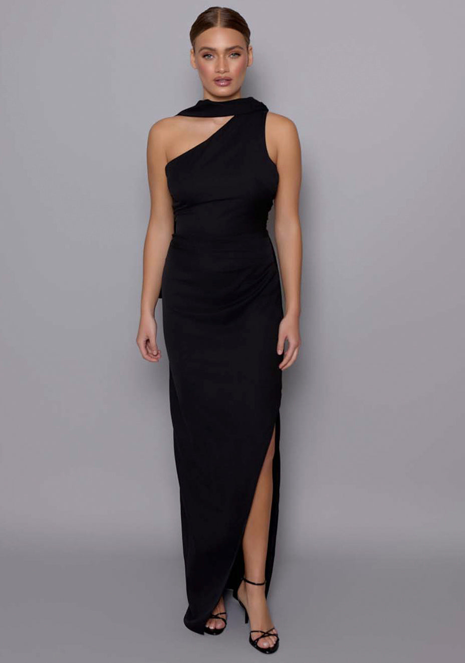 Model wearing the VSPR Drew black maxi dress with an asymmetric one-shoulder neckline, elongated shoulder drape, and side thigh split, against a grey studio backdrop.