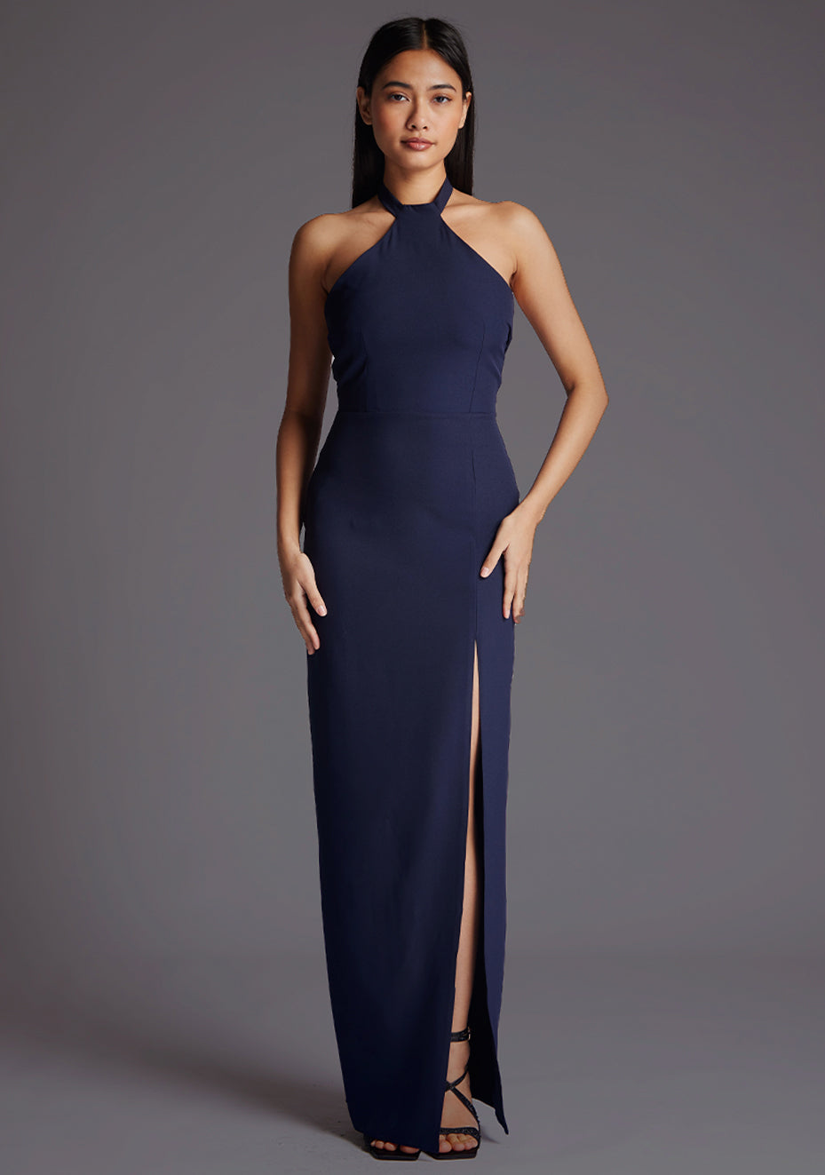 Front image of a model wearing a navy maxi dress with a  halter neck, open back, elegantly tied at the back, adorned with a chic cutout detail and front skirt split imbues a hint of allure. The design featured is the Vesper Edna Halter Neck Maxi Dress in Navy