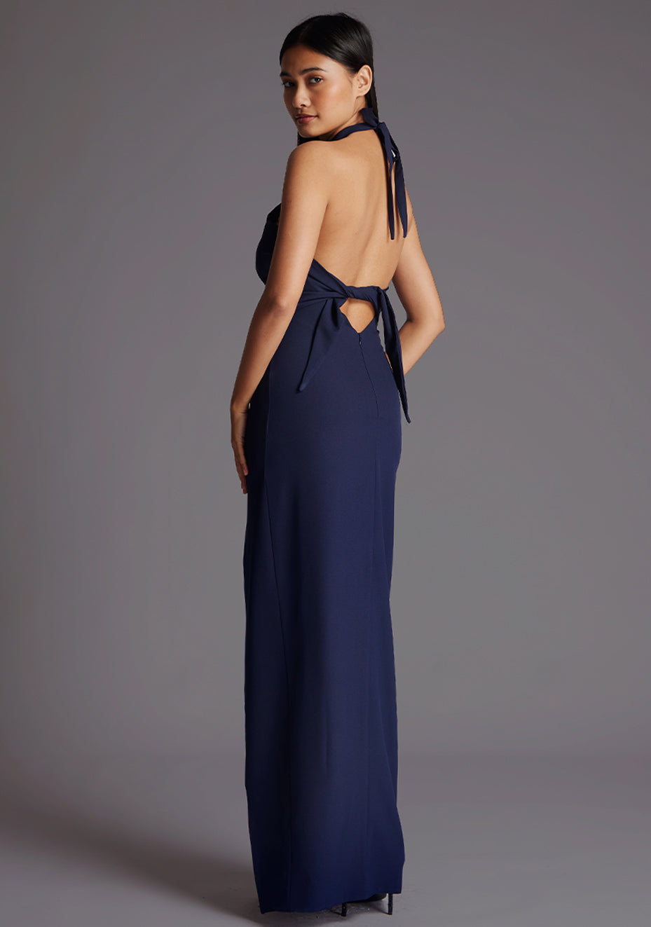 Side back quarter image of a model wearing a navy maxi dress with a  halter neck, open back, elegantly tied at the back, adorned with a chic cutout detail and front skirt split imbues a hint of allure. The design featured is the Vesper Edna Halter Neck Maxi Dress in Navy.