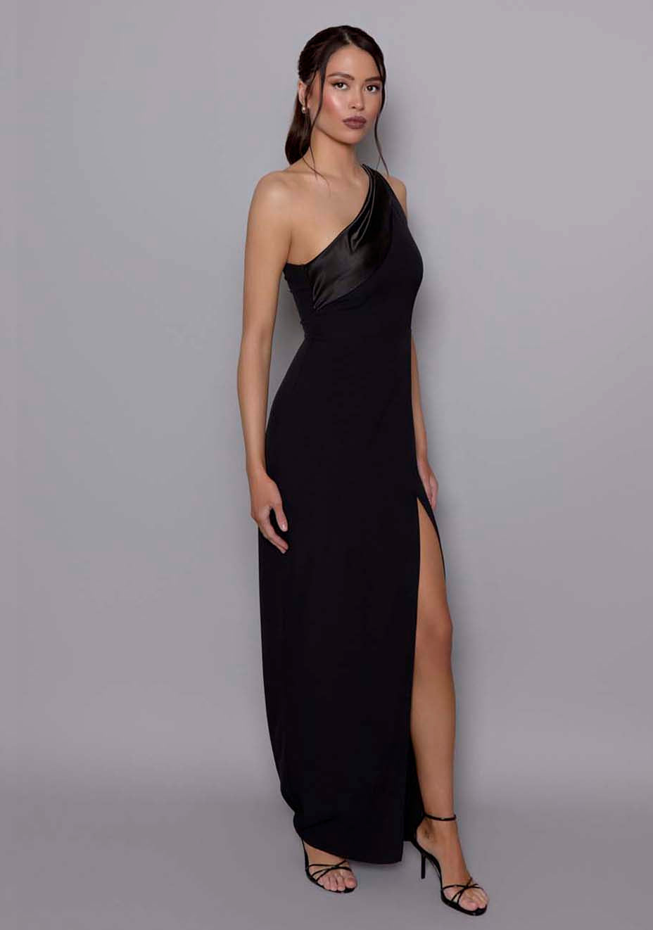 Side‑front view of the VSPR Fliss one‑shoulder black maxi dress revealing the curve‑skimming fit, satin detailing along the neckline, and the dramatic slit that elongates the leg line.