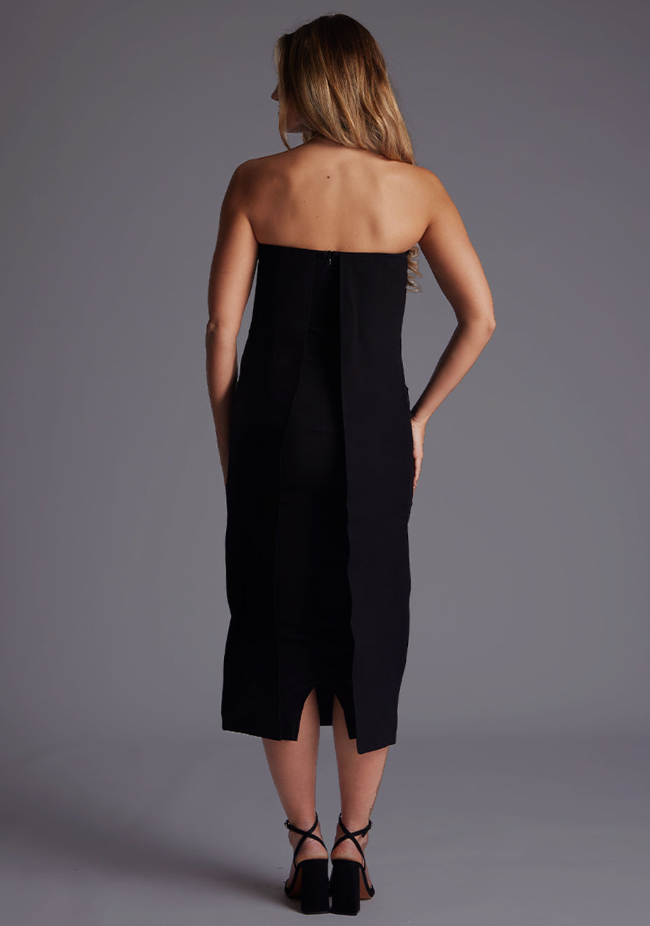 Back image of a model wearing a black midaxi dress with a featuring a straight neckline, mid-length hem and flowing fabric at beck. The design featured is the Vesper Gabriel Black Midaxi Dress