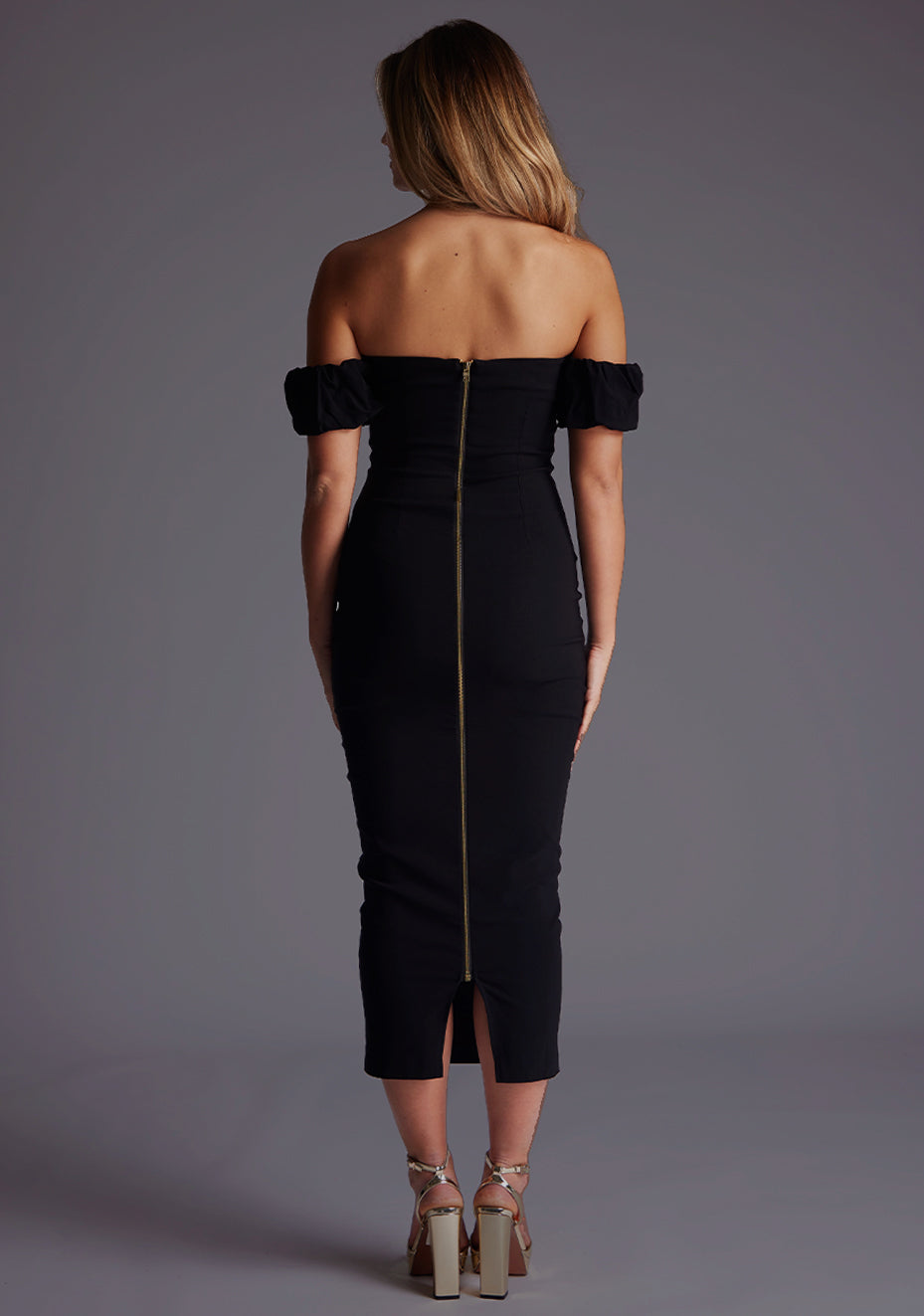 Back image of a model wearing a black bardot midaxi dress with a  bardot neckline with off shoulder puff sleeves and a statement gold centre back zip. The design featured is the Vesper Hailey Bardot Neckline Midaxi Dress in Black.