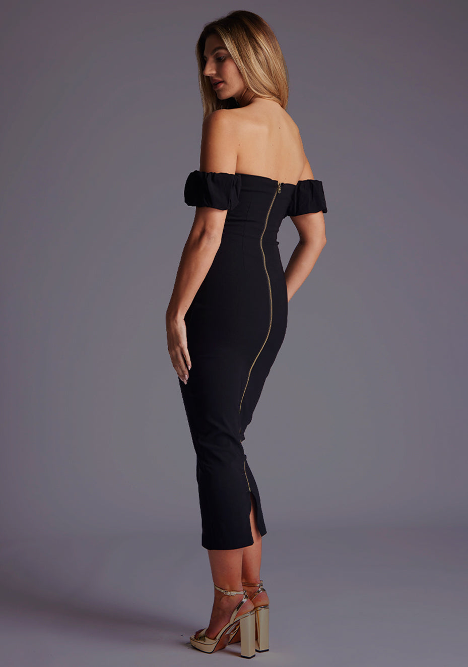 Side back image of a model wearing a black bardot midaxi dress with a  bardot neckline with off shoulder puff sleeves and a statement gold centre back zip. The design featured is the Vesper Hailey Bardot Neckline Midaxi Dress in Black.