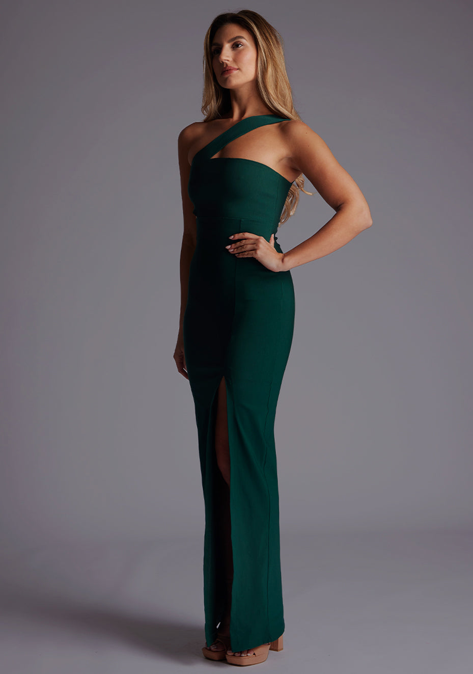 Side front image of a model wearing the Vesper Hayden Asymmetrical Maxi Dress in Forest Green, revealing the asymmetric neckline and body-contouring design.