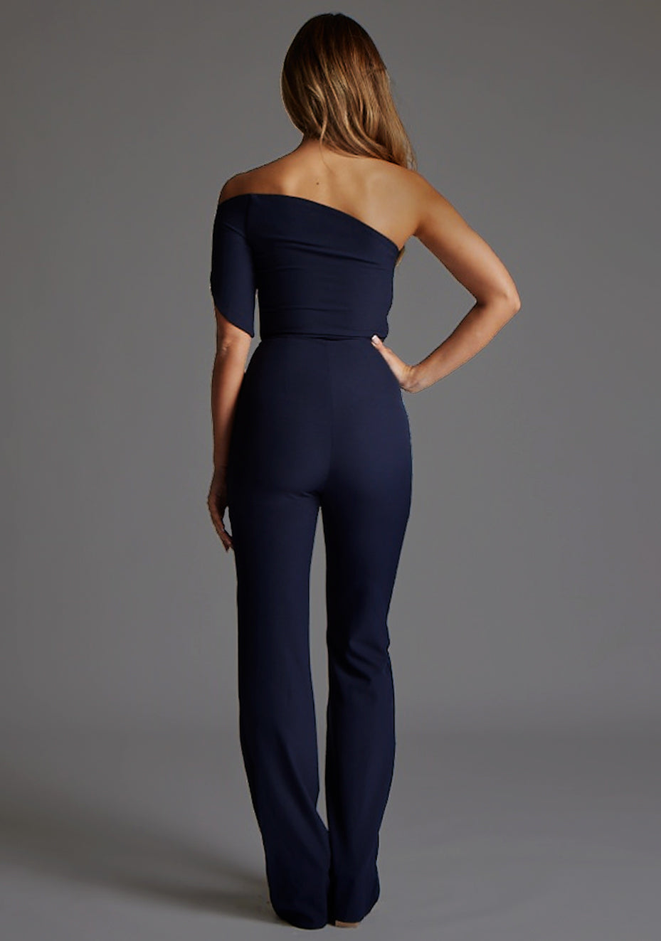 Back image of a model wearing a  navy jumpsuit with a stunning one-shoulder design and a body-hugging fit that accentuates your curves in all the right places and side zip fastening ensures a seamless look. The design featured is the Vesper Ines One Shoulder Jumpsuit in Navy.