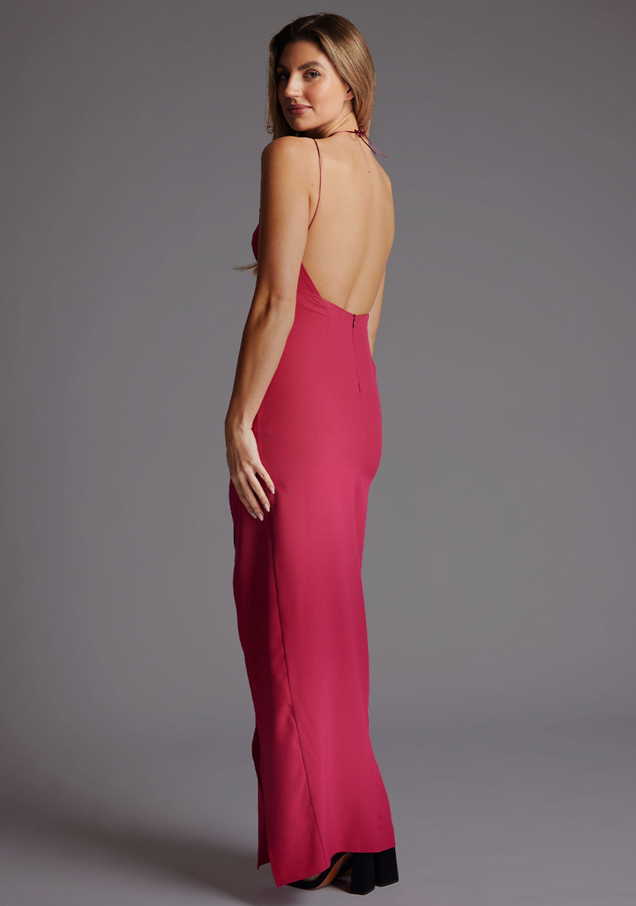 Side back image of a model wearing a raspberry strappy maxi dress with a halter neckline elegantly fastened at the back, boasting a subtle keyhole detail, revel in its backless silhouette and front skirt split. The design featured is the Vesper Ivie Strappy Open Back Maxi Dress in Raspberry.