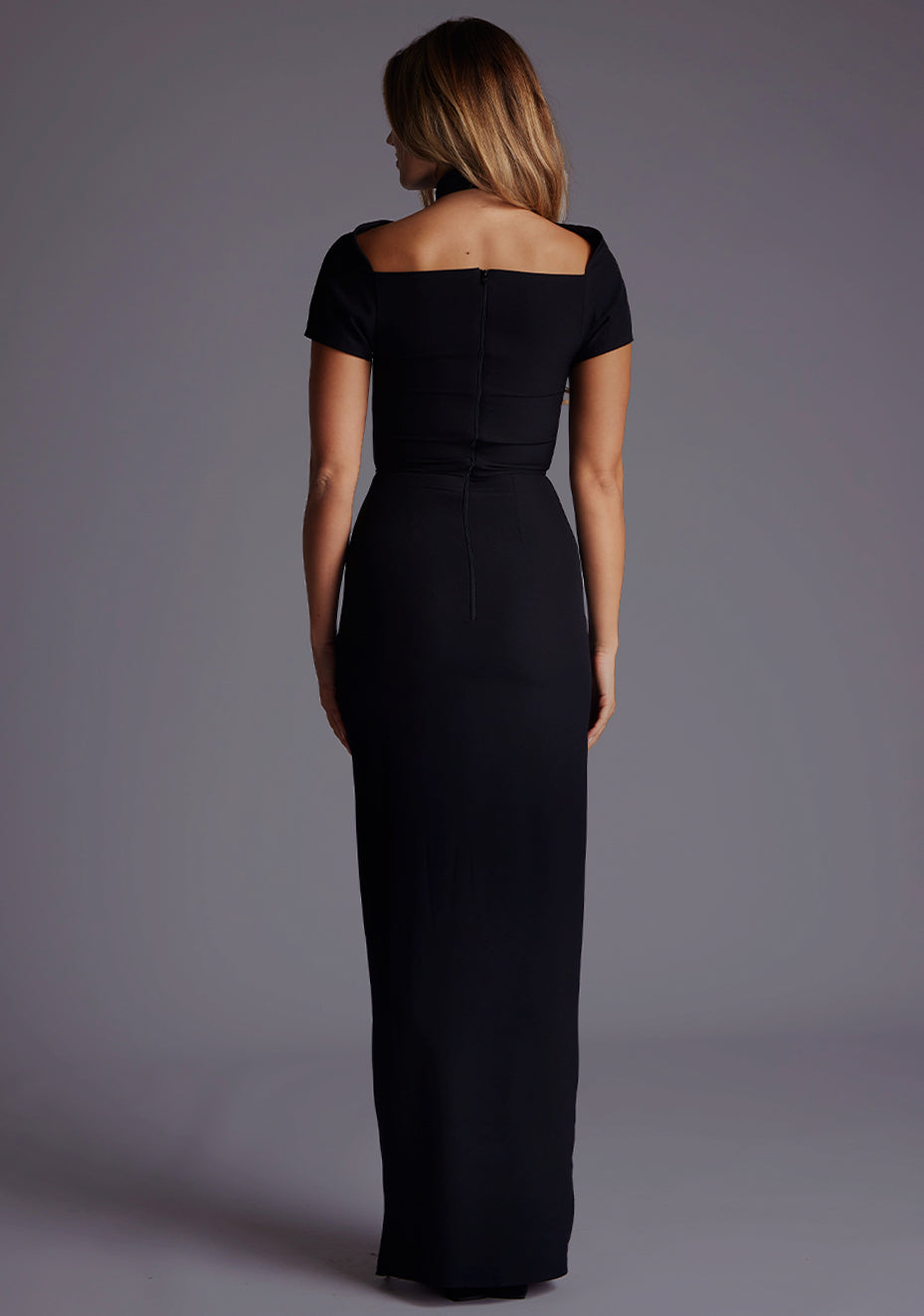 Back image of a blonde model wearing a Black Maxi Dress with a high neck, with a subtle cut-out detail, and a front skirt split. The dress is a design feature is the  Vesper Josephine High Neck Short Sleeve Maxi Dress in Black.