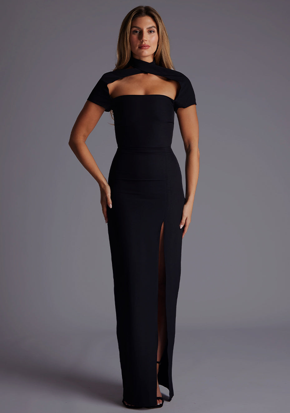 Front image of a blonde model wearing a Black Maxi Dress with a high neck, with a subtle cut-out detail, and a front skirt split. The dress is a design feature is the Vesper Josephine High Neck Short Sleeve Maxi Dress in Black.
