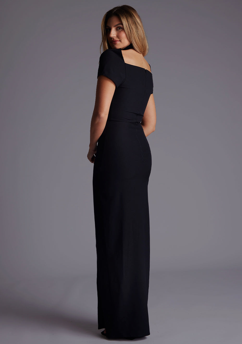 Side back image of a blonde model wearing a Black Maxi Dress with a high neck, with a subtle cut-out detail, and a front skirt split. The dress is a design feature is the  Vesper Josephine High Neck Short Sleeve Maxi Dress in Black.