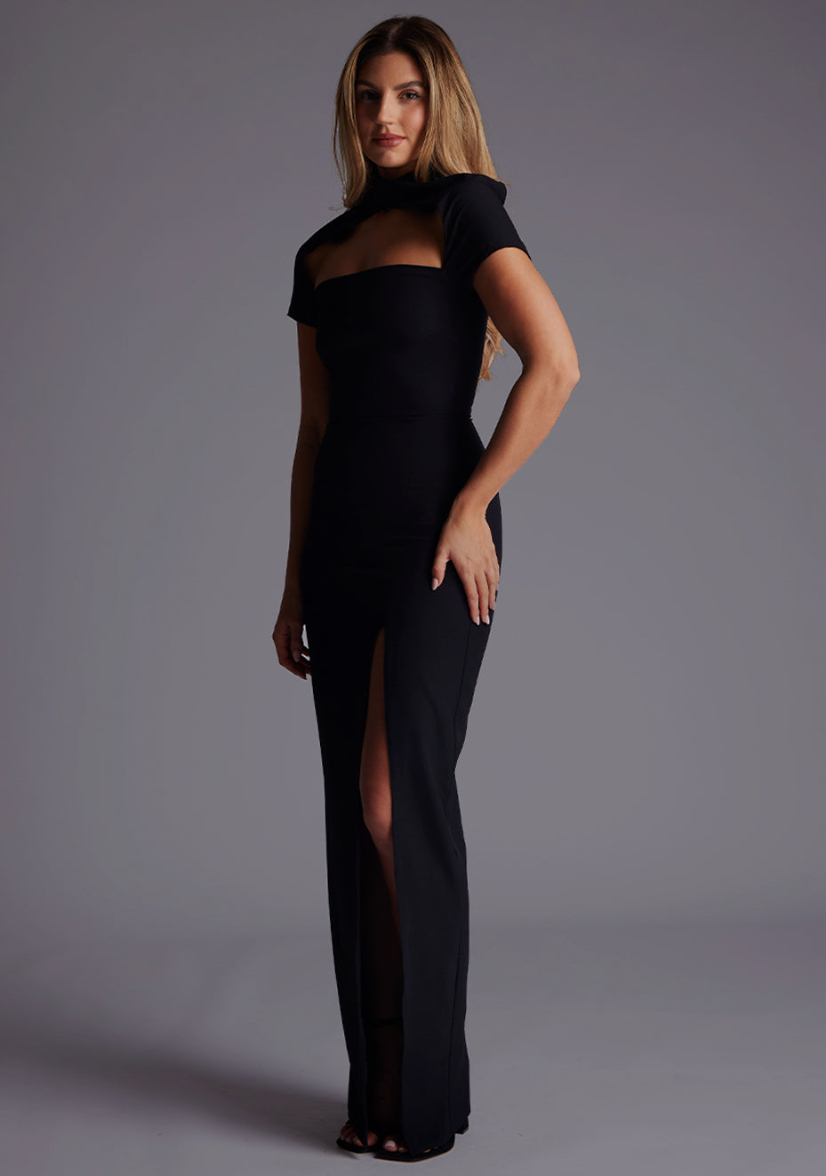 Side front image of a blonde model wearing a Black Maxi Dress with a high neck, with a subtle cut-out detail, and a front skirt split. The dress is a design feature is the  Vesper Josephine High Neck Short Sleeve Maxi Dress in Black.