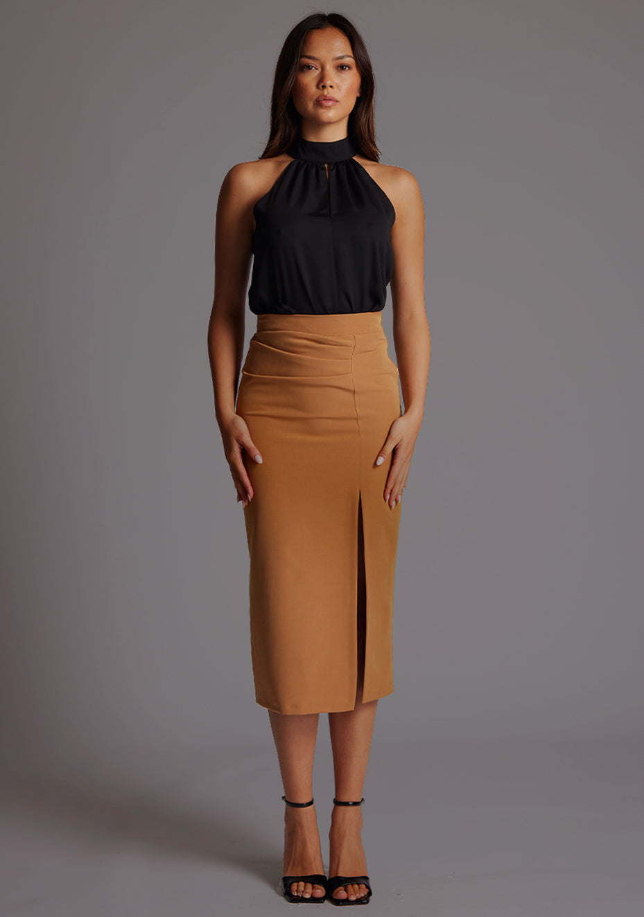 Front image of the model wearing  black top with a  subtle cut-out detail, adding a touch of modern flair to your outfit.  The design featured is the  Vesper Juniper Halter High-Neck Top in Black paired with Sherine Camel Skirt.
