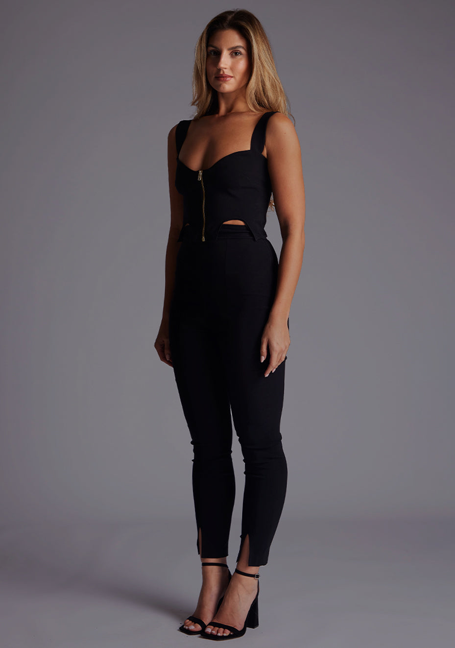 Side front image of the model wearing pine green top with a sweetheart neckline and wide straps create a flattering silhouette, while the circular cut outs at the hem add a touch of edge and statement front zip. The design featured is the Vesper Juno Sweetheart Crop Top in Black with Vesper Raven High Waist Trousers in Black.