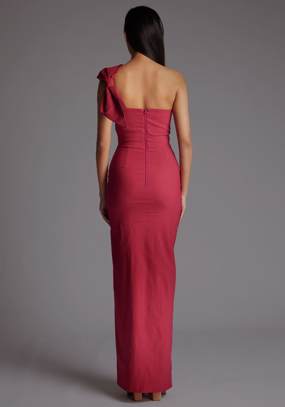 Back image of a model wearing a raspberry maxi dress, featuring an asymmetric neckline, one shoulder with statement bow detail, and a front skirt split. The dress feature is the  Vesper Keegan One Shoulder Maxi Dress in Raspberry.