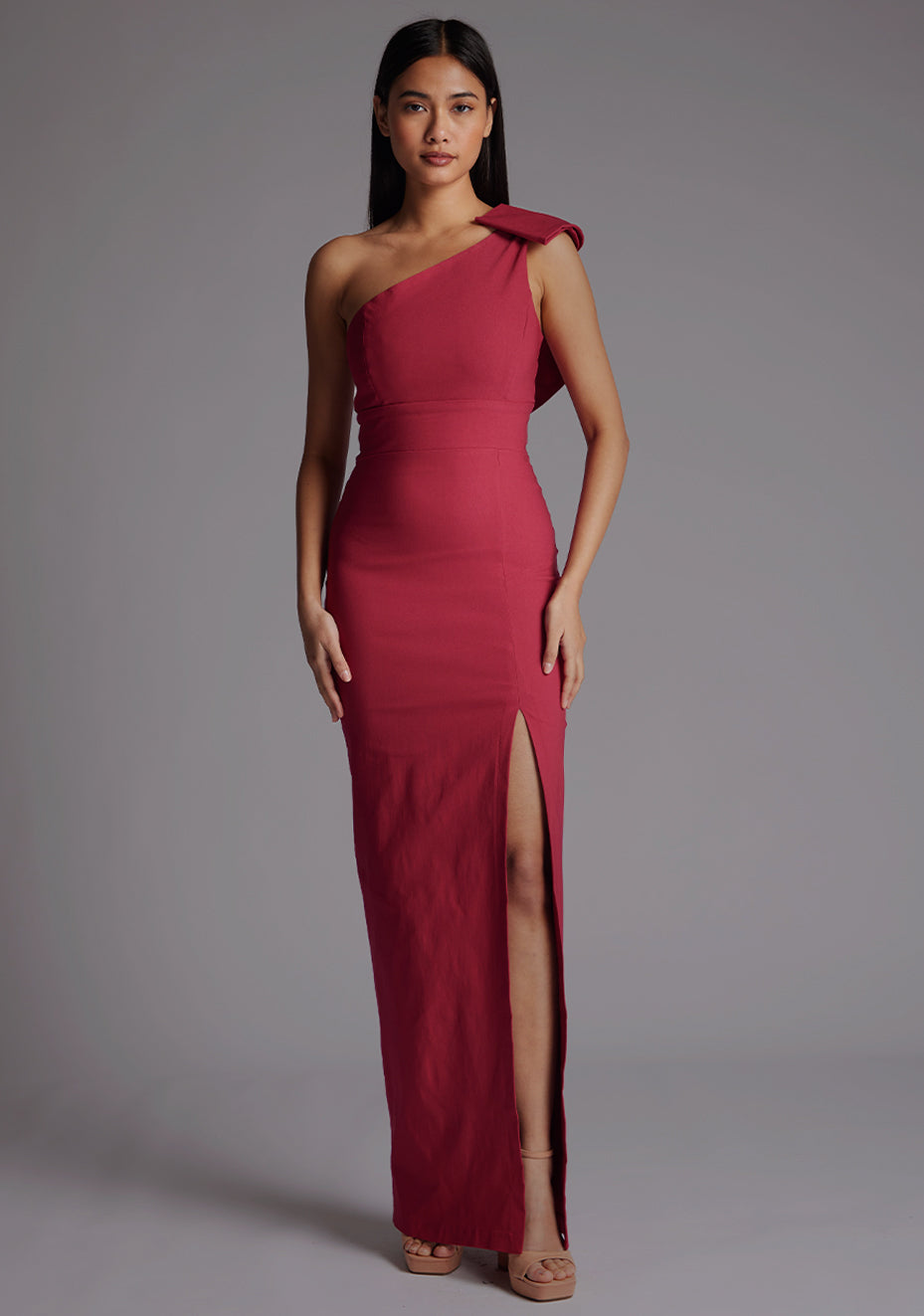 Front image of a model wearing a raspberry maxi dress, featuring an asymmetric neckline, one shoulder with statement bow detail, and a front skirt split. The dress feature is the Vesper Keegan One Shoulder Maxi Dress in Raspberry 