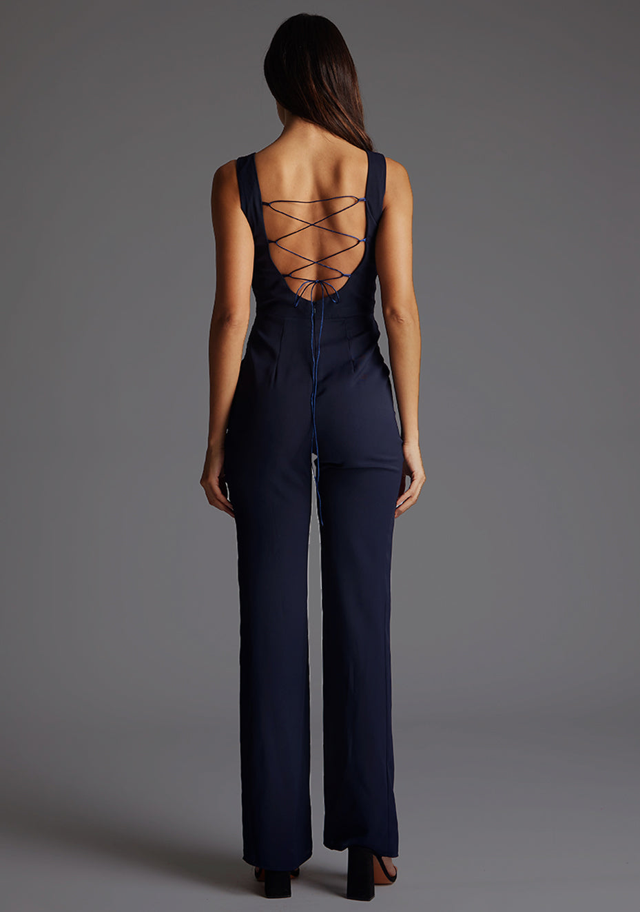 Back image of a model wearing a navy jumpsuit wit a high neckline at the front and a open back with strap detailing across the back. The design featured is the  Vesper Lilium Lace Up Back Wide Leg Jumpsuit in Navy.