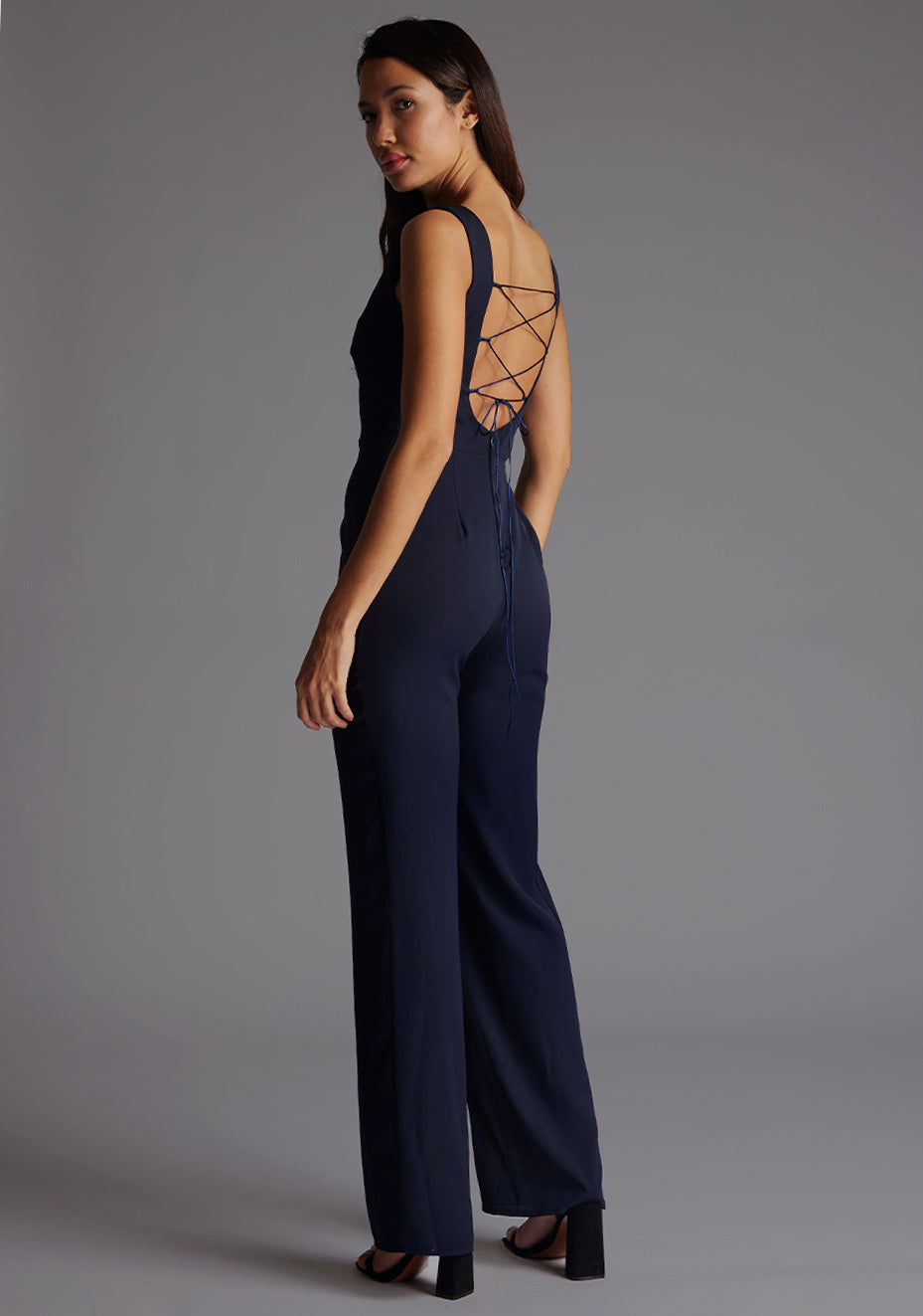 Back quarter image of a model wearing a navy jumpsuit wit a high neckline at the front and a open back with strap detailing across the back. The design featured is the  Vesper Lilium Lace Up Back Wide Leg Jumpsuit in Navy.
