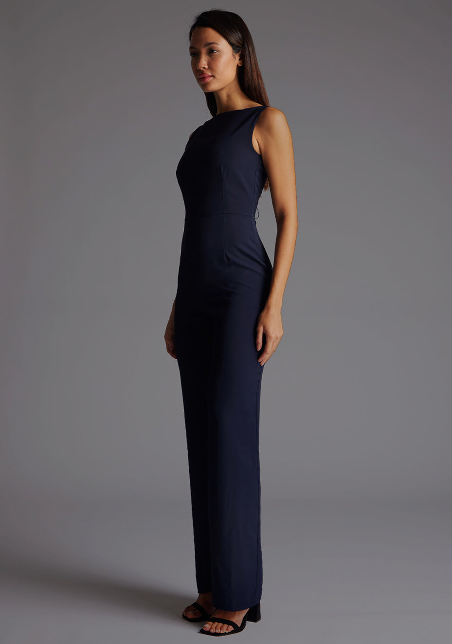 Front quarter image of a model wearing a navy jumpsuit wit a high neckline at the front and a open back with strap detailing across the back. The design featured is the  Vesper Lilium Lace Up Back Wide Leg Jumpsuit in Navy.