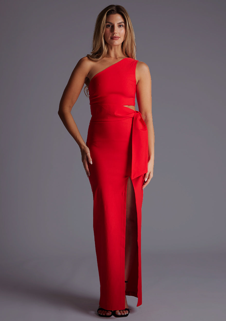 Front image of a model wearing the Vesper Lisbeth One Shoulder Maxi Dress in Red, capturing an asymmetric neckline, wrap belt waist, and front thigh split, standing against a grey studio backdrop.