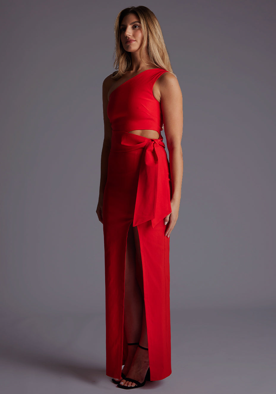 Side front image of a model wearing the Vesper Lisbeth One Shoulder Maxi Dress in Red, highlighting the wrap belt with cut-out detail and flowing skirt with front split.