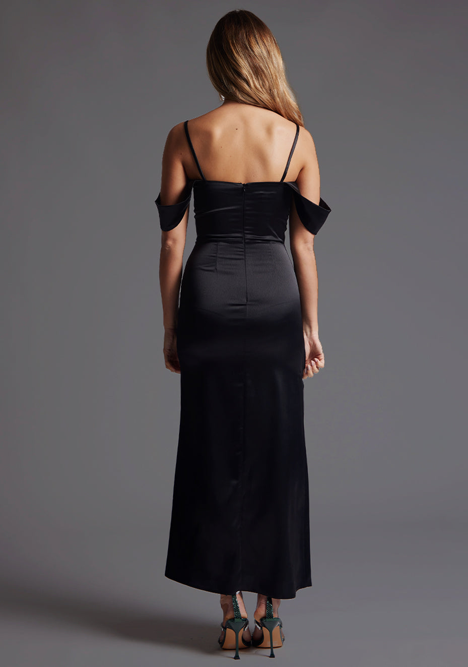 Back image of a blonde model wearing a black maxi dress with a striking black off-shoulder strap detail, beautifully complementing its slim-fit silhouette and zip closure at the back for ease and a seamless finish. The design features Vesper Luisa Bardot Satin Maxi Dress in Black.