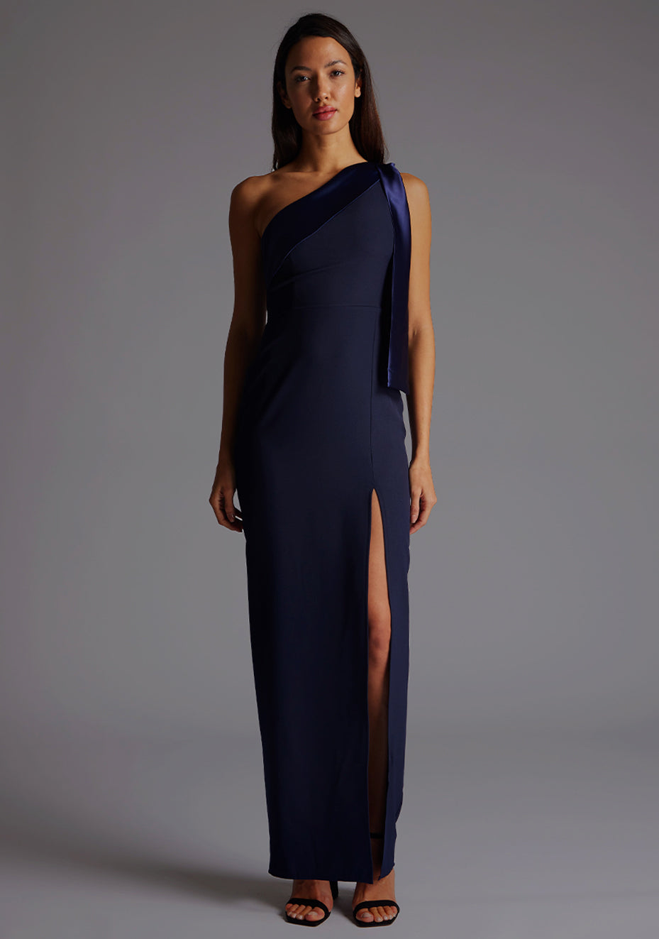 Front image of a model wearing a  One Shoulder Navy Maxi Dress with a  one shoulder contrast satin band and bow detail with a front skirt split. The design features Vesper Olympia One Shoulder Maxi Dress in Navy.