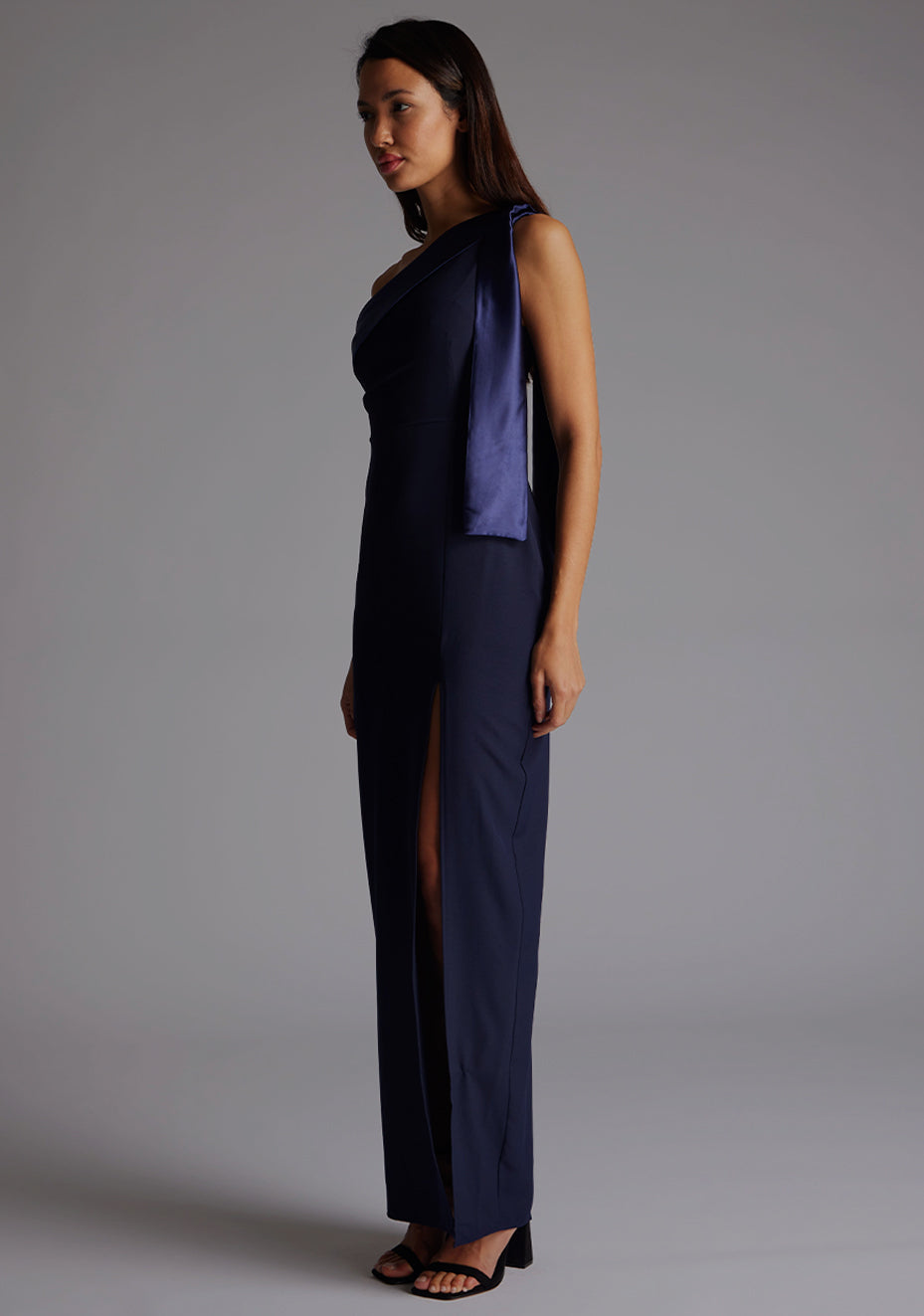 Side Front image of a model wearing a  One Shoulder Navy Maxi Dress with a  one shoulder contrast satin band and bow detail with a front skirt split. The design features Vesper Olympia One Shoulder Maxi Dress in Navy.