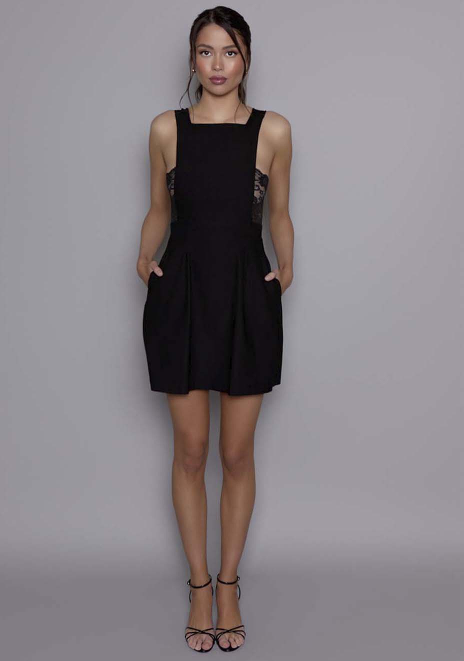 Model wearing the VSPR Noa Open Back Mini Dress in black with a squared neckline, wide straps, and a fitted bodice, standing against a gray studio backdrop.
