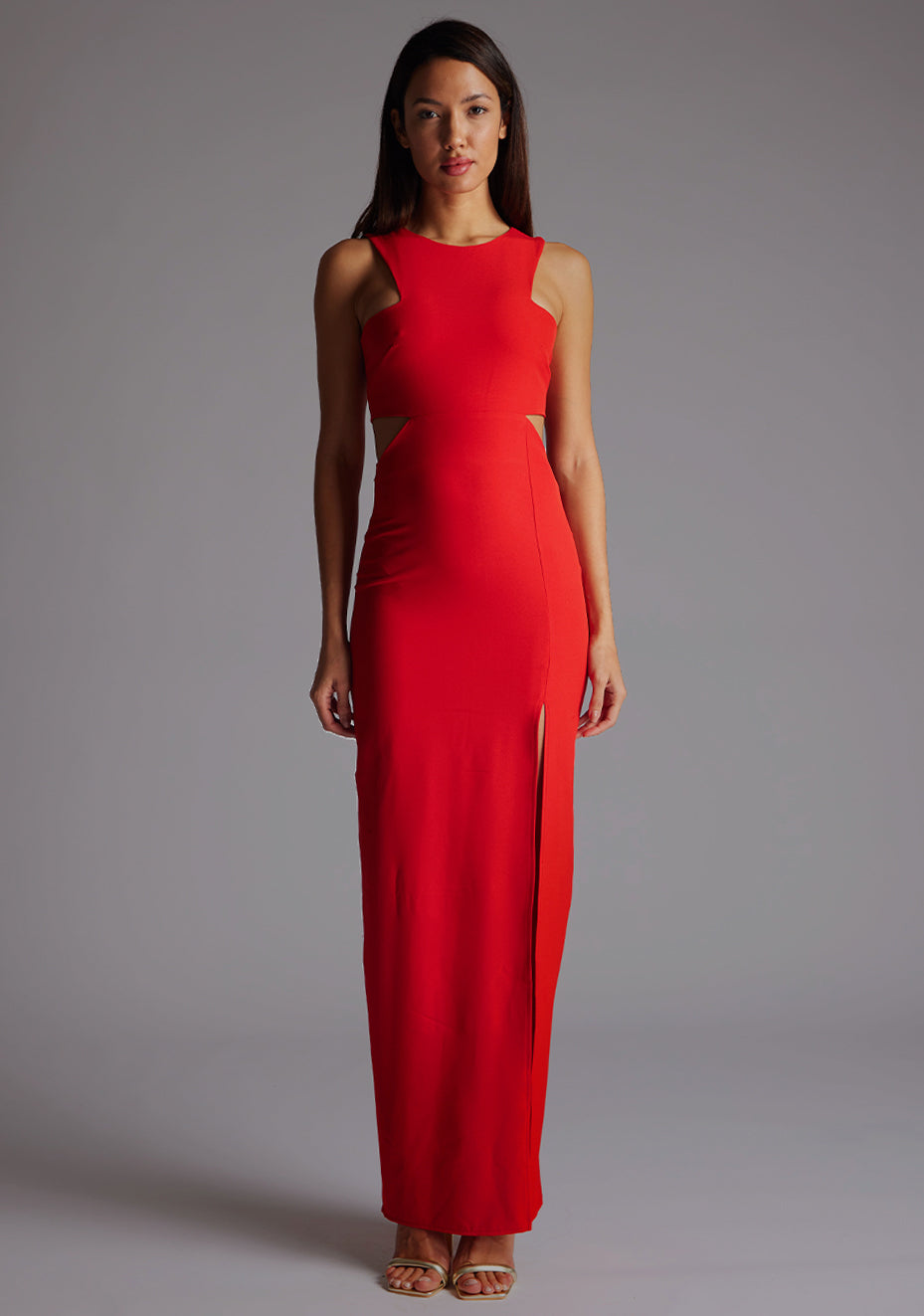 Front image of a model wearing the Vesper Paige Cut Out Maxi Dress in Red with a round neckline, side-waist cut-outs, and front split, standing against a grey studio backdrop.
