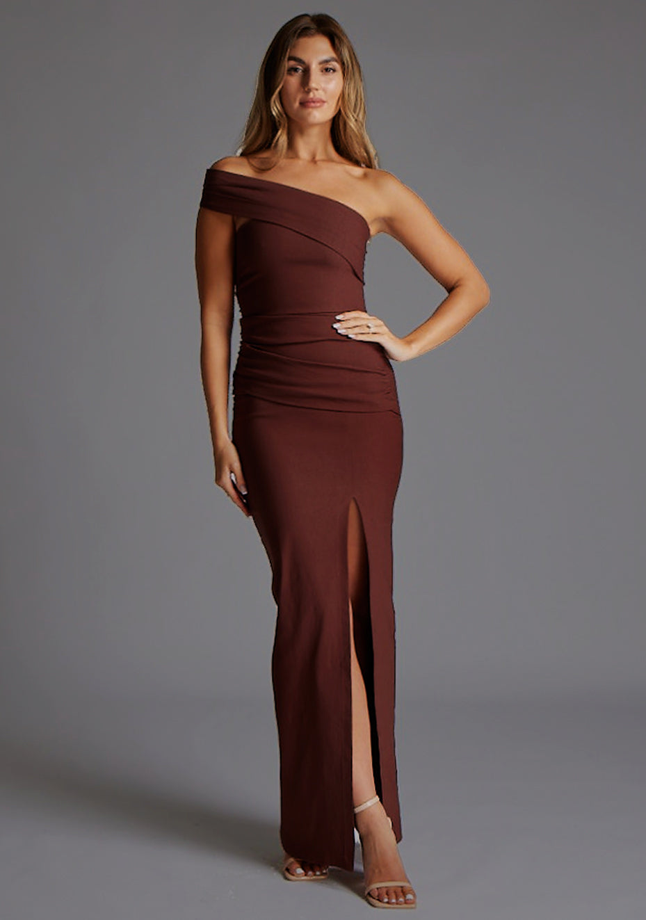 Front image of a blonde model wearing a  Chocolate Maxi Dress with an asymmetric neckline and one-sleeve design and front skirt split adds allure. The design features Vesper Pearson Bardot Ruched Maxi Dress in Chocolate.