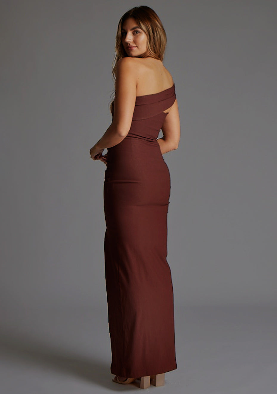 Side back image of a blonde model wearing a  Chocolate Maxi Dress with an asymmetric neckline and one-sleeve design and front skirt split adds allure. The design features Vesper Pearson Bardot Ruched Maxi Dress in Chocolate.