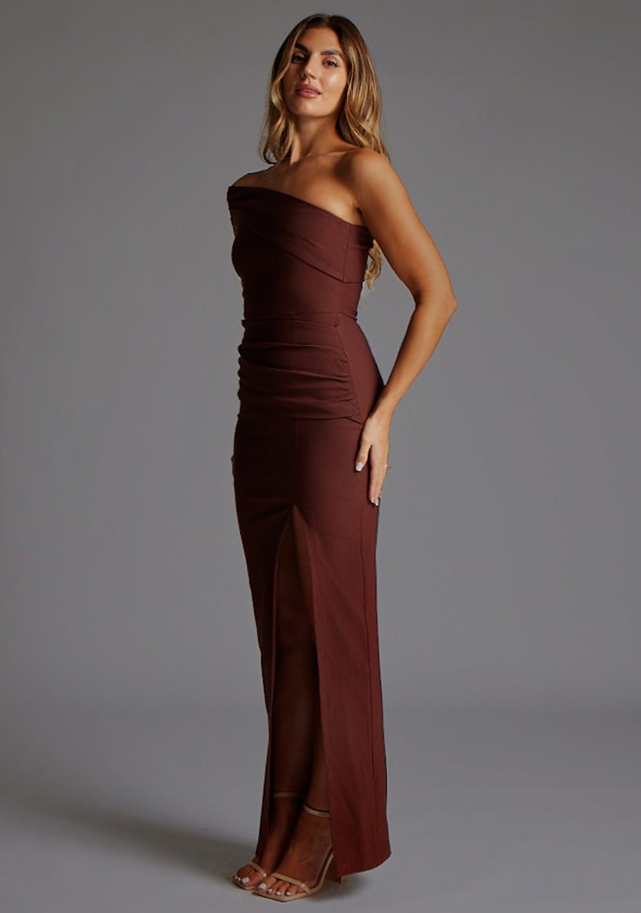 Side front image of a blonde model wearing a  Chocolate Maxi Dress with an asymmetric neckline and one-sleeve design and front skirt split adds allure. The design features Vesper Pearson Bardot Ruched Maxi Dress in Chocolate.