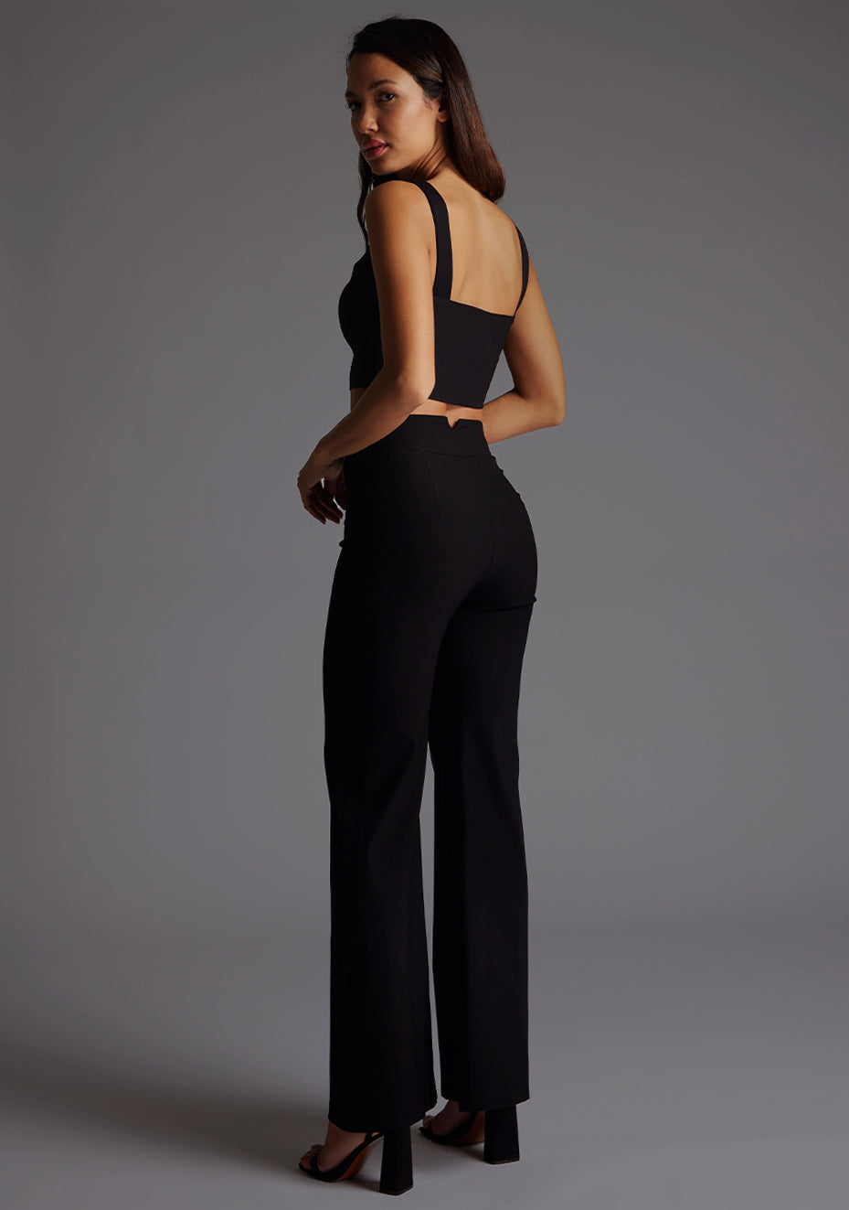 Side back image of a model wearing a black crop top with a sweetheart neckline and thick straps. The design featured is the Vesper Sweetheart Neckline Crop Top in Black pair with Vesper June Wide Leg Trousers in Black.