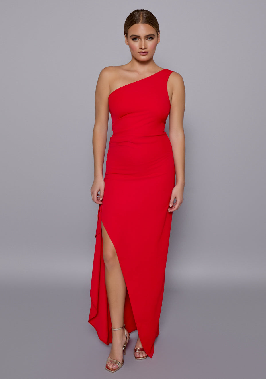 Model wearing the VSPR Stella Red One-Shoulder Maxi Dress with asymmetrical neckline and thigh-high front slit, standing against a neutral studio backdrop.