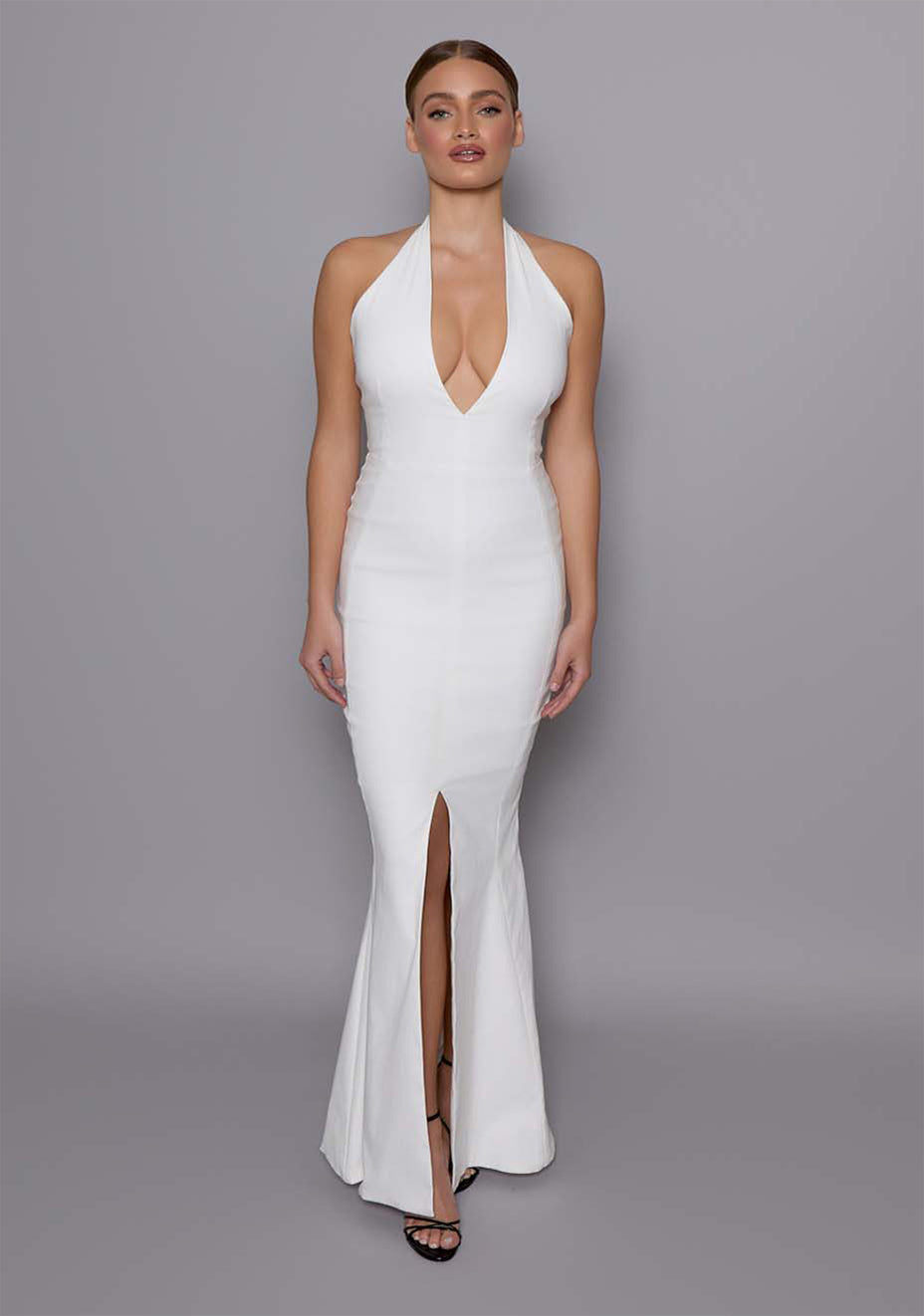 Model wearing the VSPR Zara Ivory Fishtail Maxi Dress with a plunging halter neckline, contoured bodice, and a thigh-high front slit, standing against a gray studio backdrop.