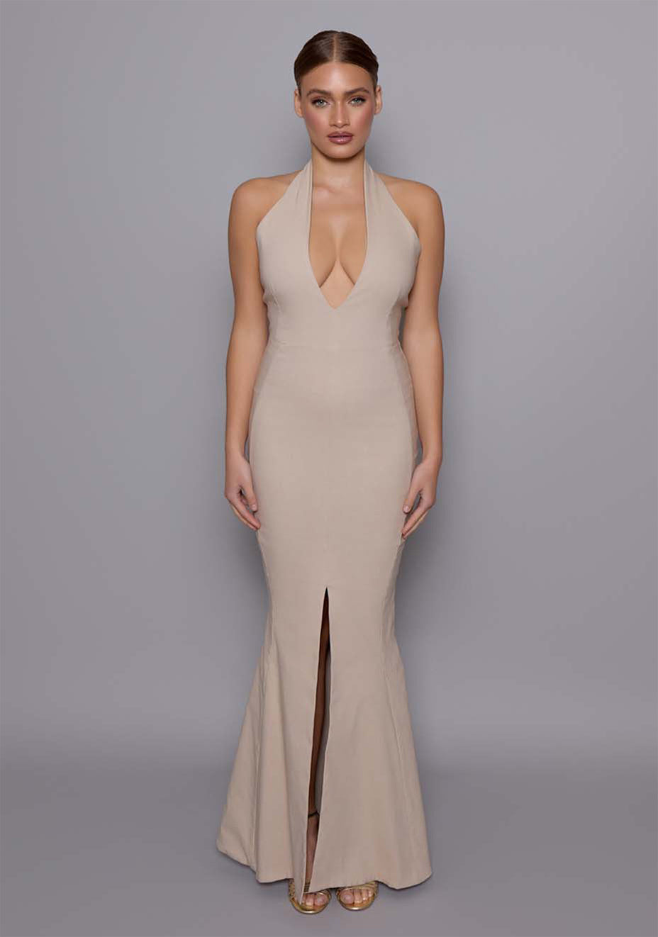 Model wearing the VSPR Zara Fishtail Maxi Dress in Stone, featuring a plunging halter neckline and contoured bodice, with a sophisticated stone hue, standing against a gray studio backdrop.