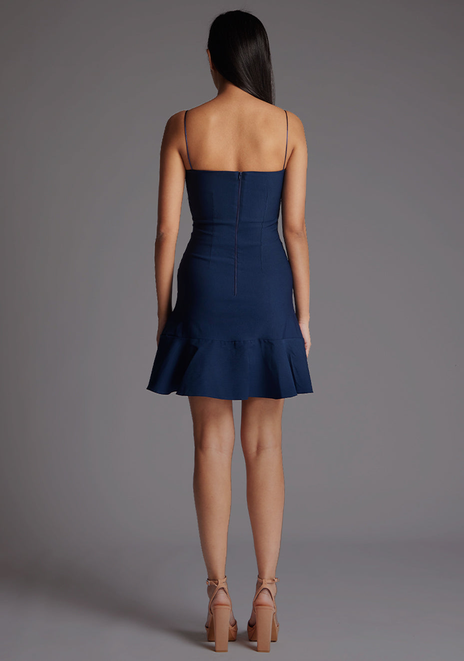 Back image of a model wearing a navy mini dress with a straight neckline, thin straps, a fitted bodice, and flared ruffle hem with invisible centre back zip. The design featured is the Vesper Brenda Navy Mini Dress.