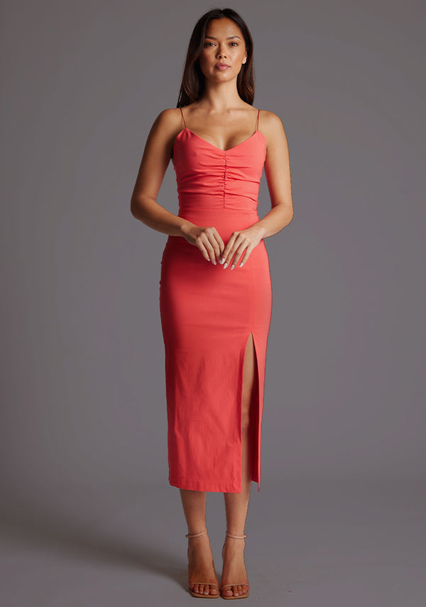 Front image of a model wearing a coral midaxi dress with slender straps, a graceful V-neckline, and a captivating front skirt split. The design featured is the Vesper Bronwen Coral Midaxi Dress.