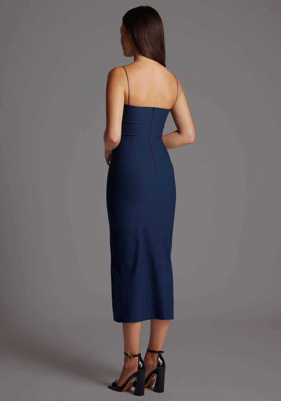 Back quarter image of a model wearing a navy midaxi dress with a timeless design boasts a subtle sweetheart neckline and sophisticated front cut-outs. The graceful front skirt split adds allure. The design featured is the Vesper Kendall Navy Midaxi Dress
