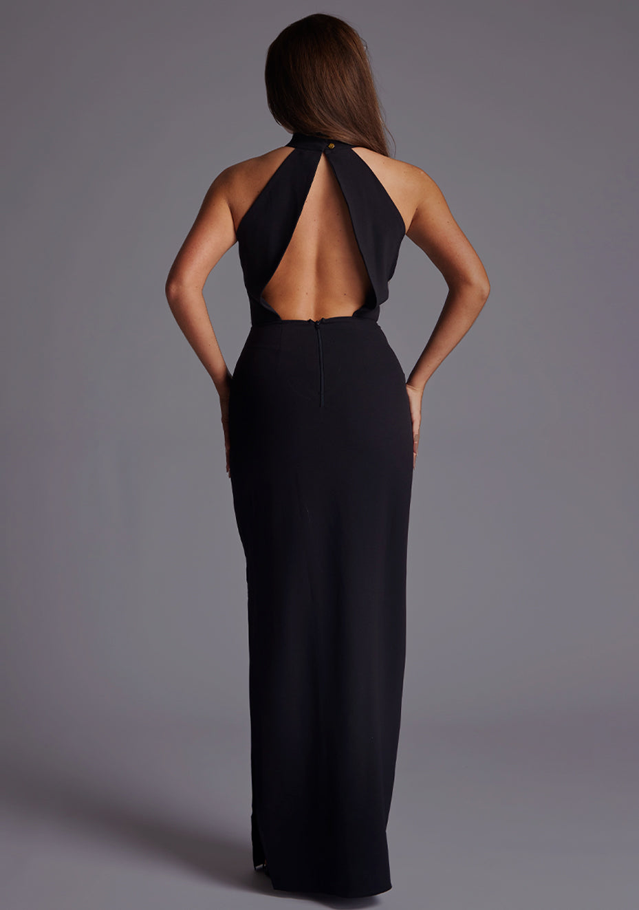Back image of a model wearing a black cut out maxi dress with a elegant maxi dress features a split at the left side, open back and front bodice keyhole detail. This number fastens at the back of the neck with a buttoned collar and centre back zip detail. The design featured is the Vesper Kennedy Black Cut Out Maxi Dress