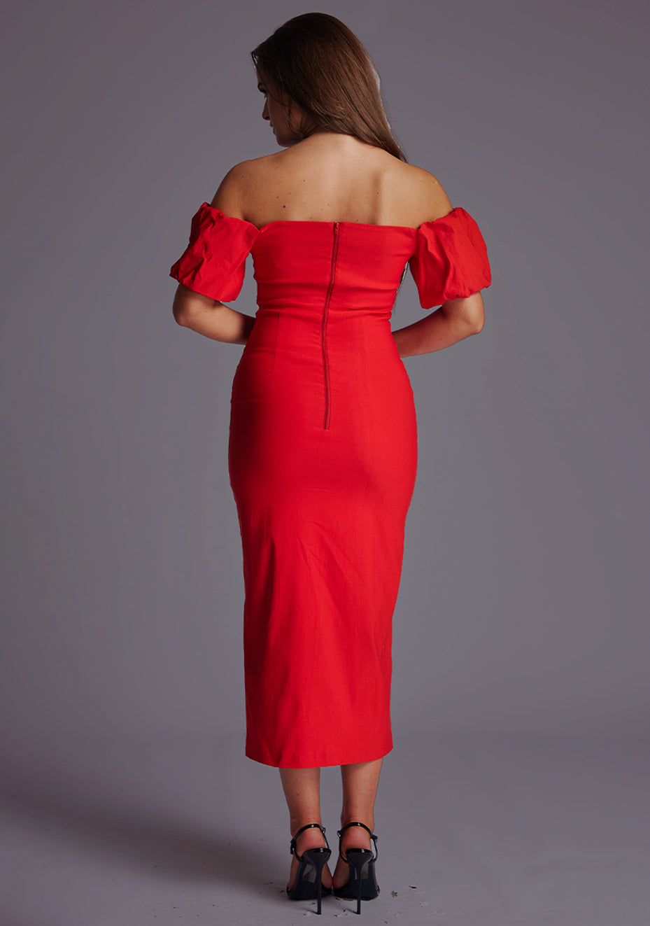 Back image of a model wearing a red midaxi dress with a off shoulder design with sweetheart neckline, puffy bands at the top of arms and front skirt split. The design featured is the Vesper Lottie Red Midaxi Dress