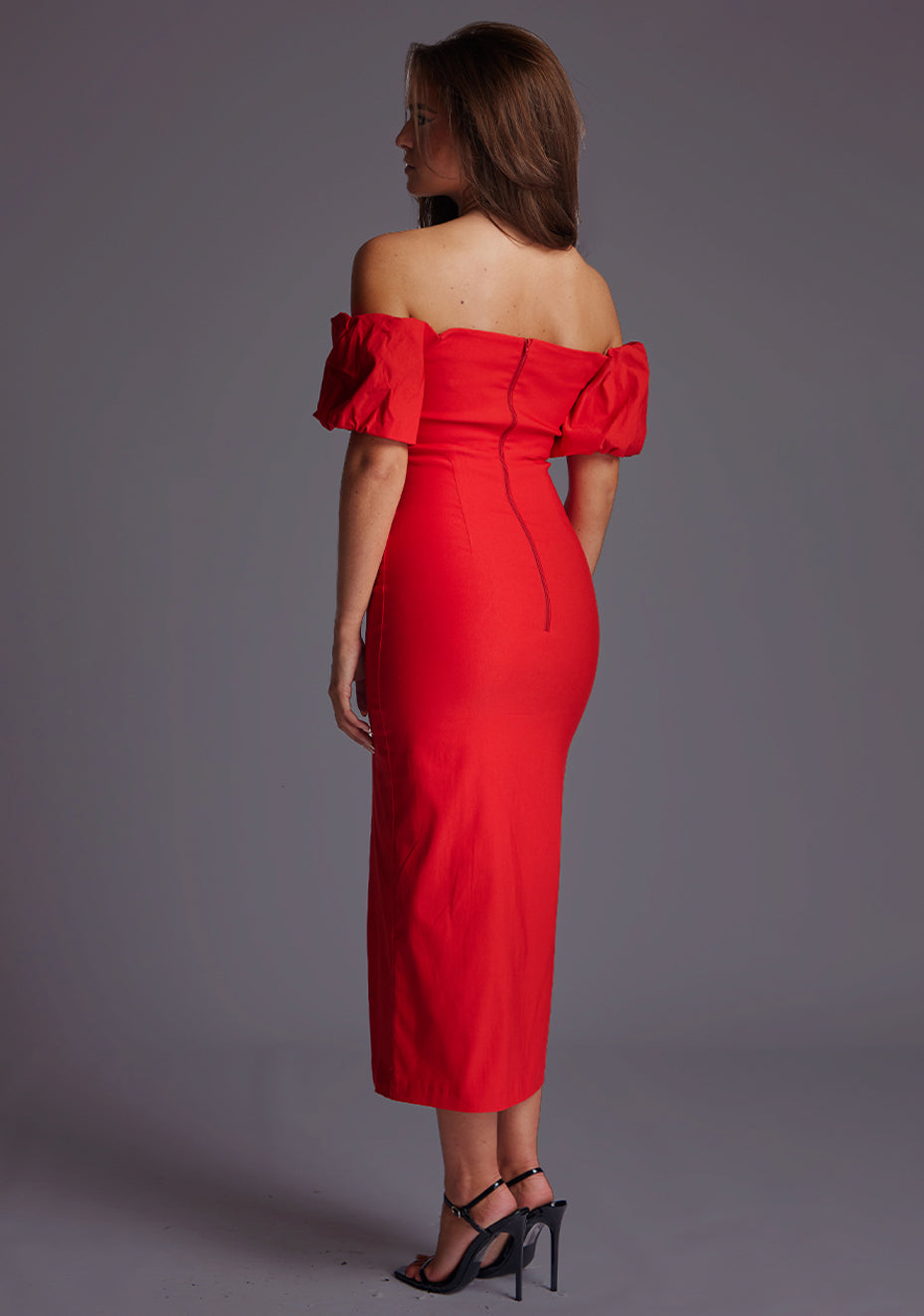 Back quarter image of a model wearing a red midaxi dress with a off shoulder design with sweetheart neckline, puffy bands at the top of arms and front skirt split. The design featured is the Vesper Lottie Red Midaxi Dress
