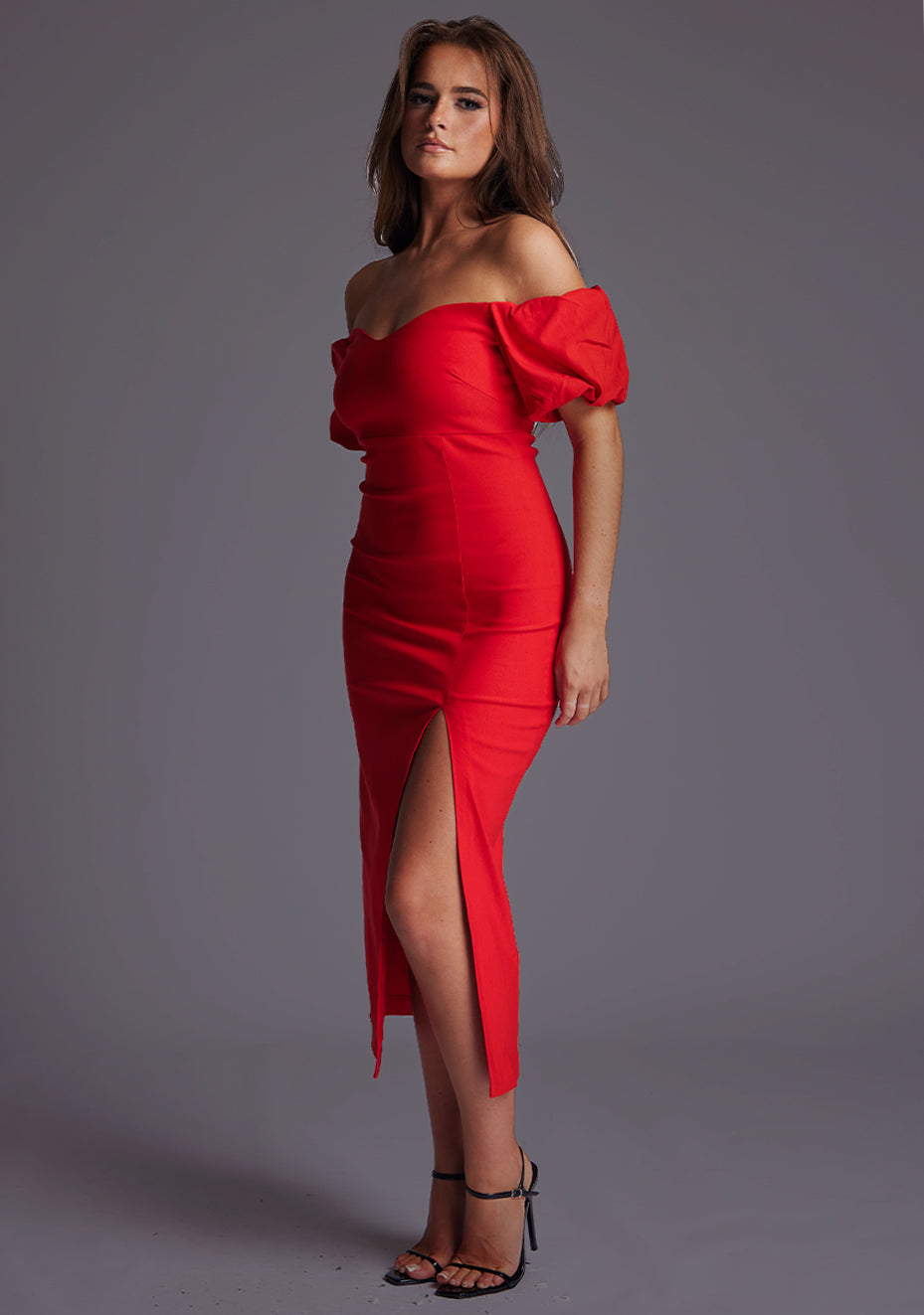 Front quarter image of a model wearing a red midaxi dress with a off shoulder design with sweetheart neckline, puffy bands at the top of arms and front skirt split. The design featured is the Vesper Lottie Red Midaxi Dress