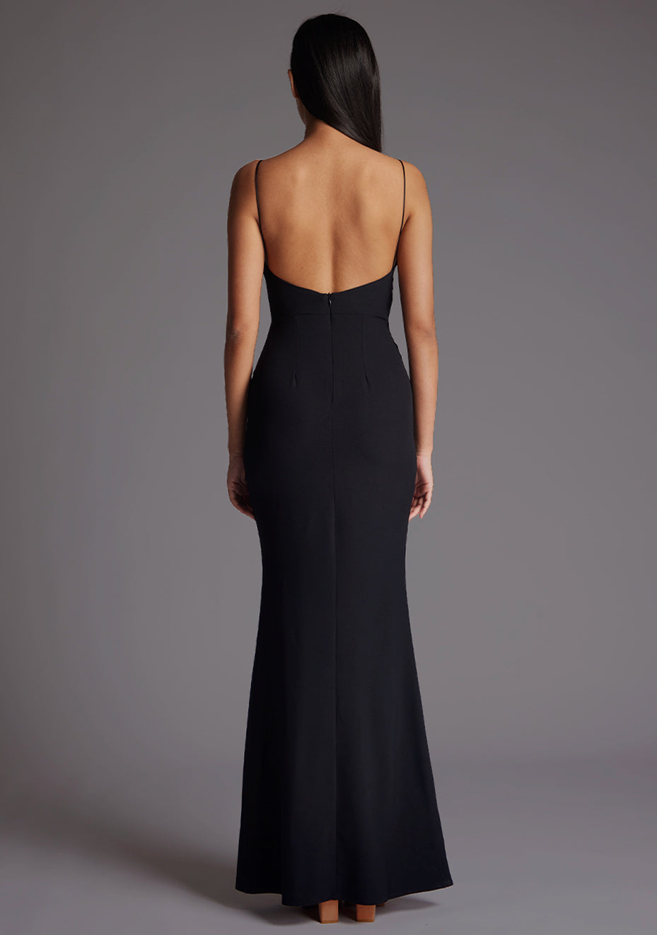 Back iimage of a model wearing a black maxi dress with a v-neckline, thin straps with twist front detail at bust, low back and subtle flair at hem. The design featured is the Vesper Marlowe Black Maxi Dress