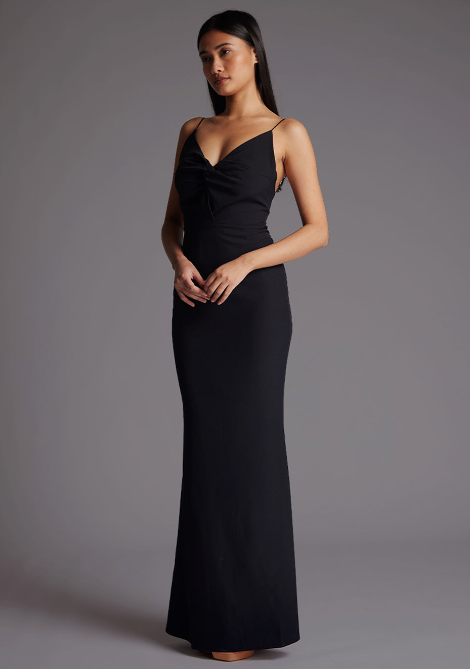 Front quarter image of a model wearing a black maxi dress with a v-neckline, thin straps with twist front detail at bust, low back and subtle flair at hem. The design featured is the Vesper Marlowe Black Maxi Dress