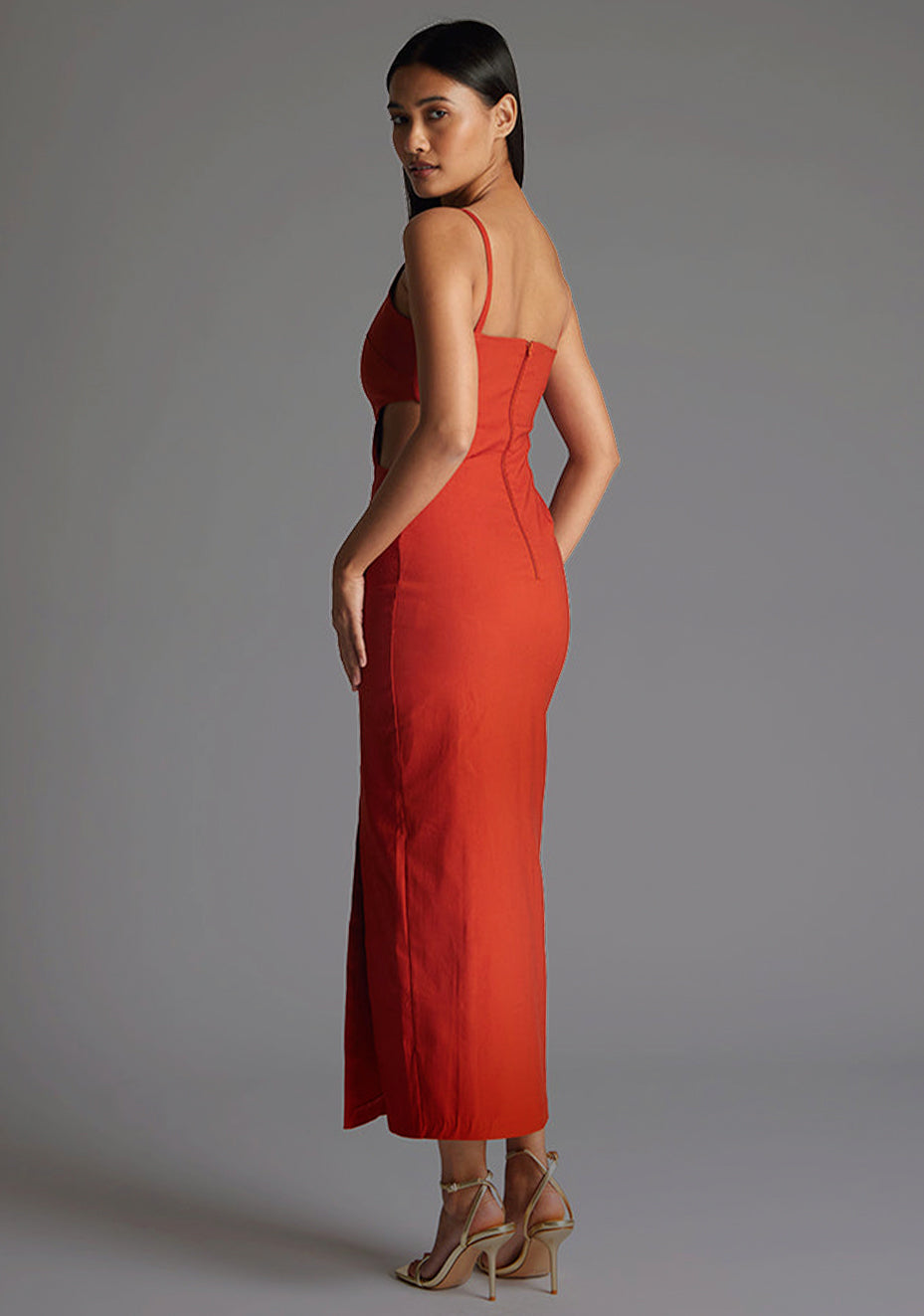 Back quarter image of the model wearing a rust midaxi dress with a scoop neckline, bodycon silhouette, side cut out and front skirt split . The design featured is the Vesper Maryam Rust Midaxi Dress.
