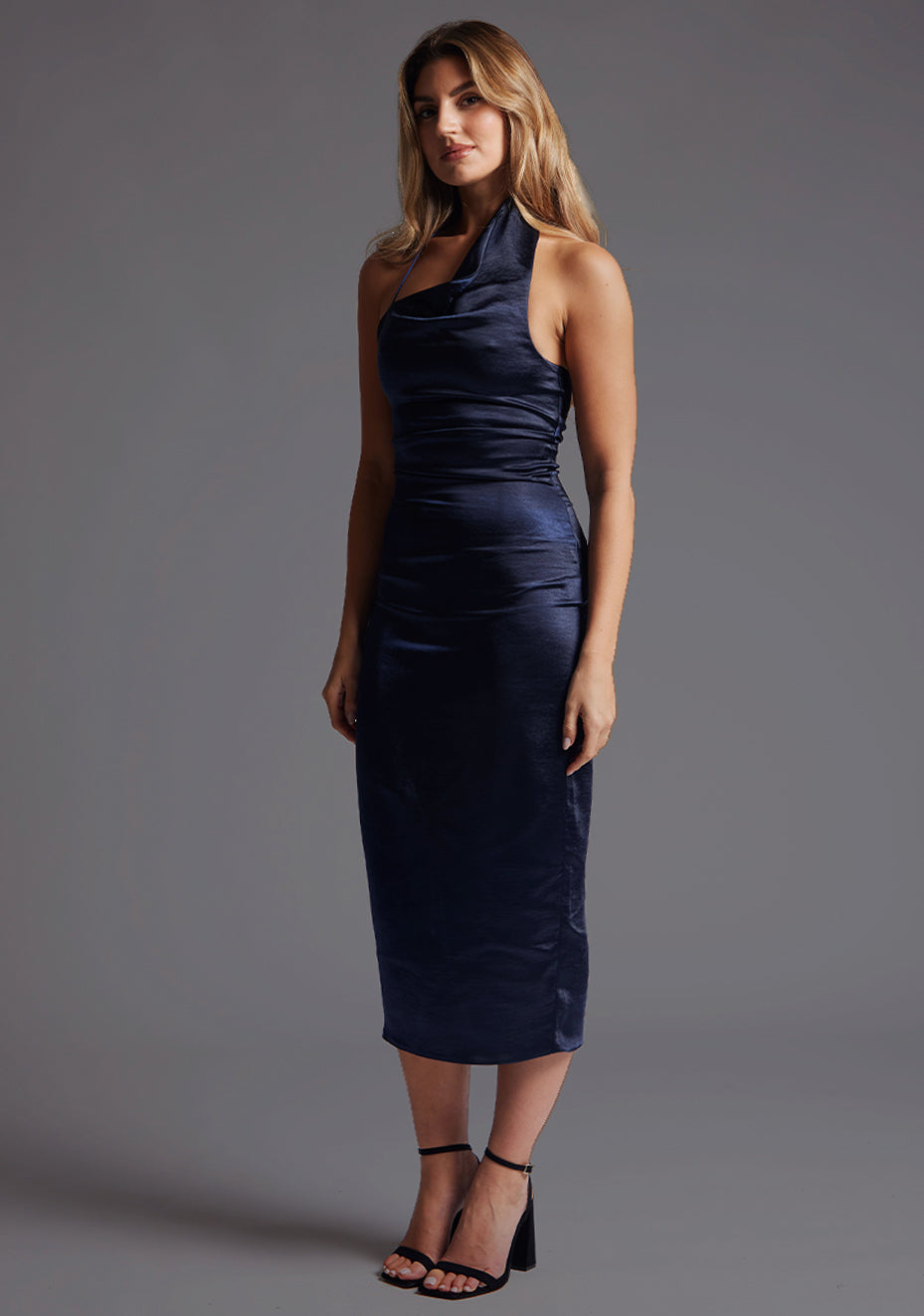 Front quarter image of a blonde model wearing a navy midaxi dress with a asymmetric neckline with one-shoulder design, captivating cowl design and slim fit detail. The design featured is the Vesper Mason Navy Midaxi Dress.