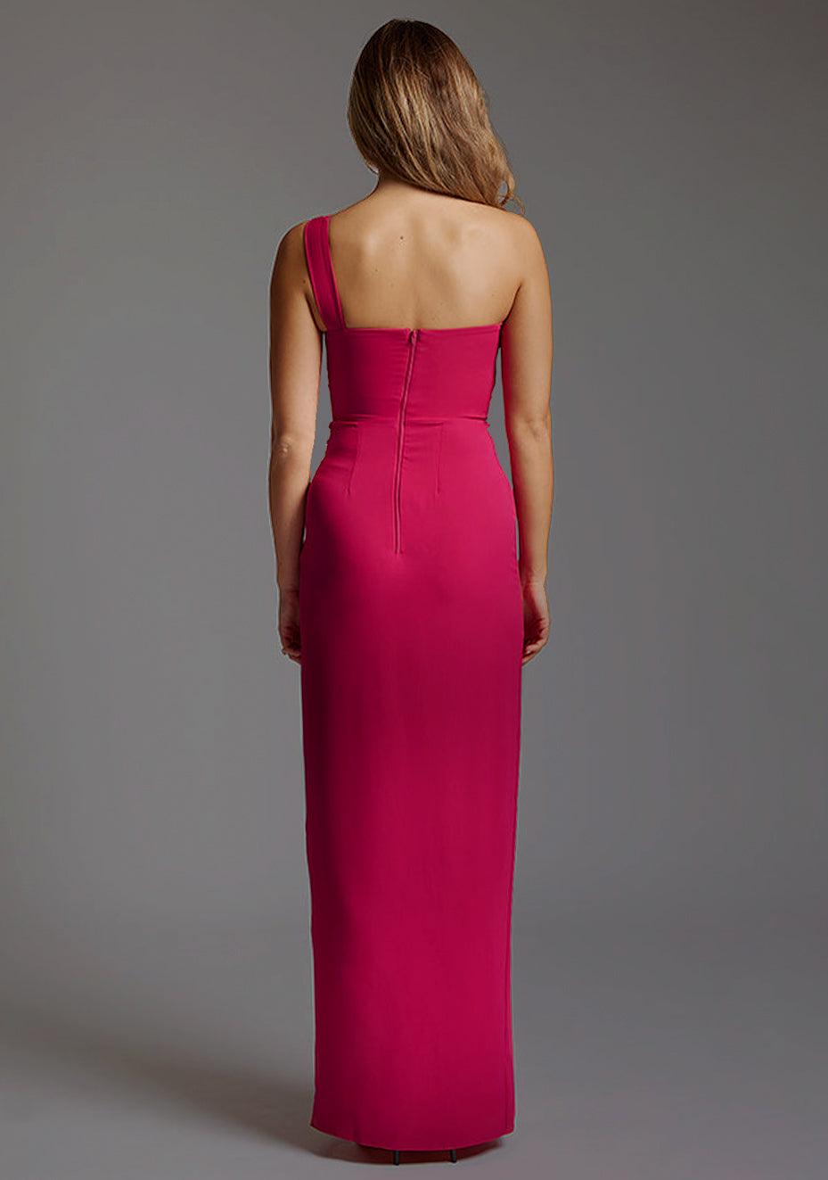 Back image of a blonde model wearing a ccerise maxi dress with a asymmetric neckline, front skirt split and beautiful cerise hue. The design featured is the Vesper Olivette Cerise Maxi Dress