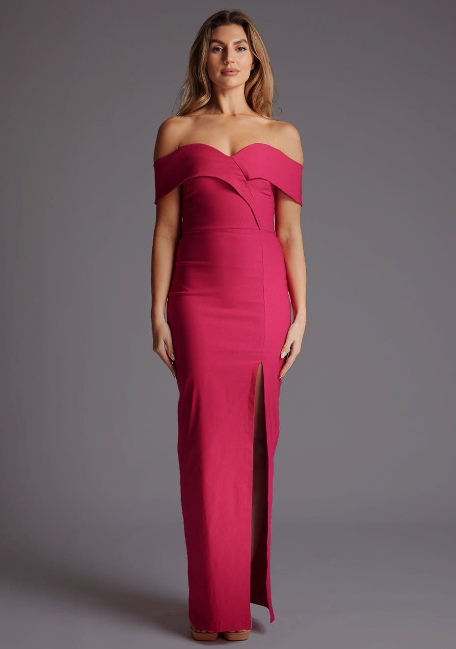 Front image of a blonde model wearing a ccerise maxi dress with a asymmetric neckline, front skirt split and beautiful cerise hue. The design featured is the Vesper Olivette Cerise Maxi Dress.