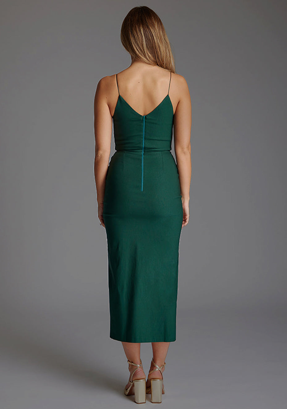 Back image of blonde model wearing a forest green midaxi dress with a plunge neckline, delicate thin straps for a feminine touch, and front split that adds a modern edge.  The design featured is the Vesper Palmer Forest Green Midaxi Dress